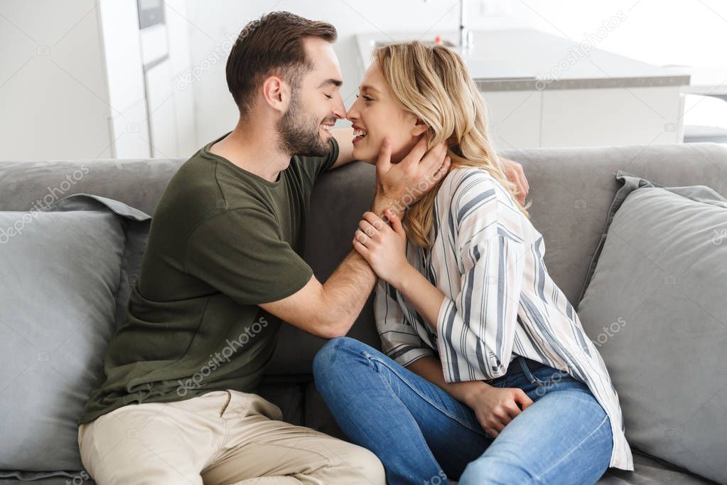 Smiling young loving couple indoors at home sitting on sofa hugging kissing.