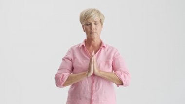 Sakin sarışın yaşlı kadın gözlerini açma ve meditasyon gri arka plan izole üzerinde elleri bir arada tutarak çıkmak