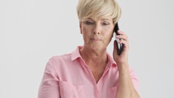 スマートフォンで電話を取り 灰色の背景の上に誰かと意見の相違を持つ深刻なブロンドの老婦人のトリミングされたビュー — ストック動画