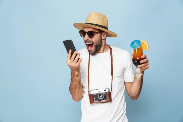 Emocionado joven turista posando con cámara usando teléfono móvil bebiendo cóctel aislado sobre fondo de pared azul . — Foto de Stock