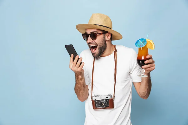 Opgewonden jonge man toerist poseren met camera met behulp van mobiele telefoon drinken cocktail geïsoleerd over blauwe muur achtergrond. — Stockfoto