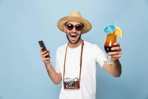 Emocionado joven turista posando con cámara usando teléfono móvil bebiendo cóctel aislado sobre fondo de pared azul . — Foto de Stock