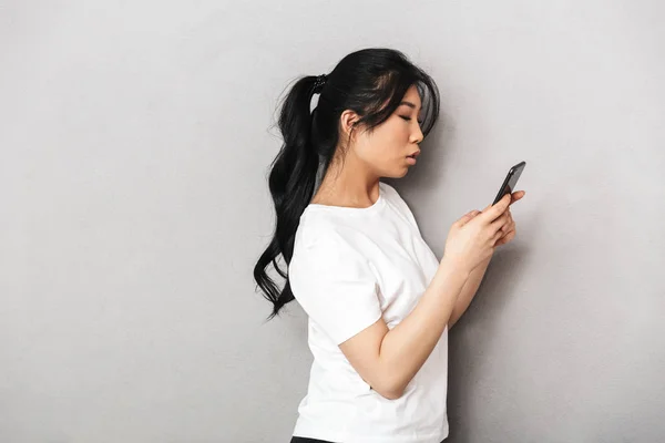 Ασίας όμορφη νεαρή γυναίκα που παρουσιάζουν απομονωμένη πάνω από γκρι τοίχο φόντο χρησιμοποιώντας το κινητό τηλέφωνο. — Φωτογραφία Αρχείου