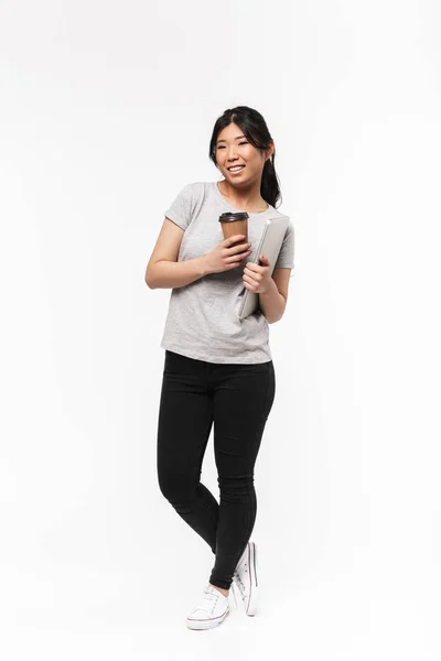 Ασίας όμορφη νεαρή γυναίκα που θέτουν απομονωμένα πάνω από λευκό τοίχο φόντο πόσιμο καφέ κρατώντας φορητό υπολογιστή. — Φωτογραφία Αρχείου