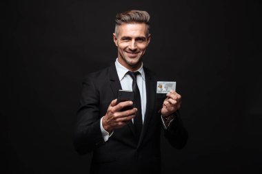 Cep telefonu ve kredi kartı tutan resmi takım elbise giymiş gülümseyen traşsız işadamı portresi