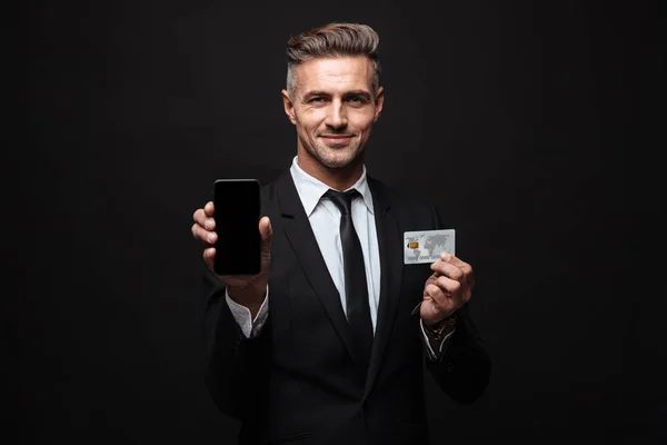 Портрет усміхненого впевненого бізнесмена, одягненого в офіційний костюм, що тримає мобільний телефон і кредитну картку — стокове фото