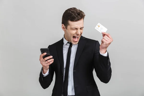 Imagen del empresario morena encantado en traje formal gritando mientras sostiene el teléfono celular y la tarjeta de crédito — Foto de Stock