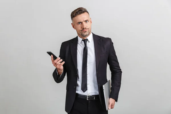 Πορτρέτο του νευρικός προβληματισμένος ενήλικος επιχειρηματίας στο γραφείο κοστούμι κρατώντας smartphone και φορητό υπολογιστή, ενώ στέκεται — Φωτογραφία Αρχείου
