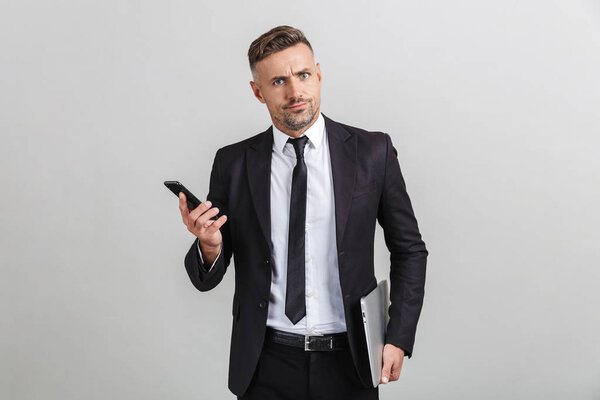 Portrait of uptight puzzled adult businessman in office suit holding smartphone and laptop while standing