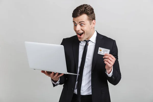 Imagem de empresário morena surpreso em terno formal surpreendente ao segurar laptop e cartão de crédito — Fotografia de Stock