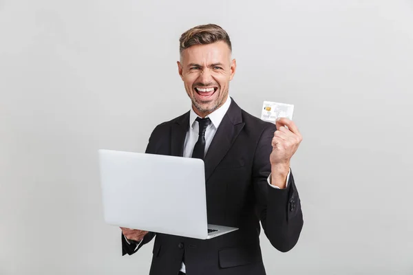 Imagen de empresario adulto positivo en traje formal regocijándose mientras sostiene el ordenador portátil y la tarjeta de crédito — Foto de Stock