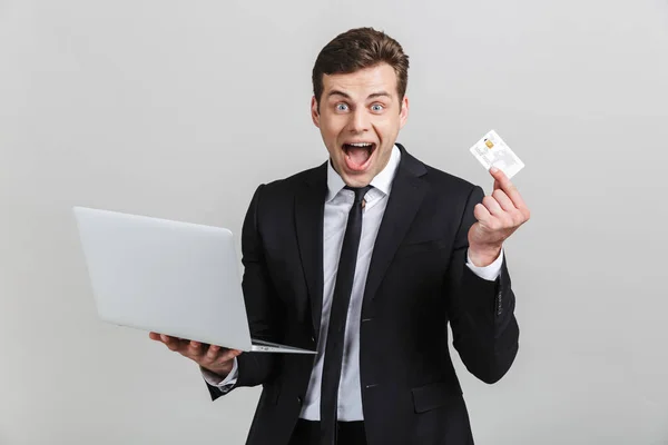 Imagen de hombre de negocios morena extática en traje formal sorprendente mientras sostiene el ordenador portátil y la tarjeta de crédito — Foto de Stock