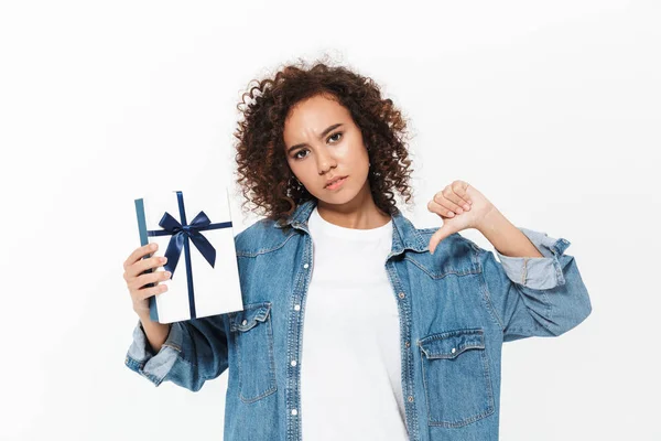 Schöne unzufriedene junge Afrikanerin posiert isoliert über weißem Wandhintergrund mit Geschenk-Box. — Stockfoto