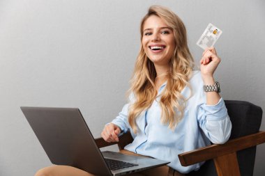 Mutlu genç güzel sarışın iş kadını dizüstü bilgisayar kullanarak kredi kartı tutarak izole gri duvar arka plan poz.