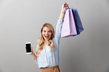Mutlu heyecanlı genç güzel sarışın iş kadını alışveriş çantaları tutarak izole gri duvar arka plan poz.