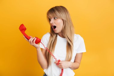 Şok genç sarışın kadın telefonla konuşurken beyaz rahat t-shirt giymiş sarı duvar arka plan üzerinde izole poz.