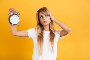 Ciddi genç sarışın kadın düşünme sarı duvar arka plan üzerinde beyaz rahat t-shirt tutan çalar saat giymiş izole poz.