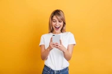 Mutlu duygusal genç sarışın kadın cep telefonu kullanarak beyaz rahat t-shirt giymiş sarı duvar arka plan üzerinde izole poz.