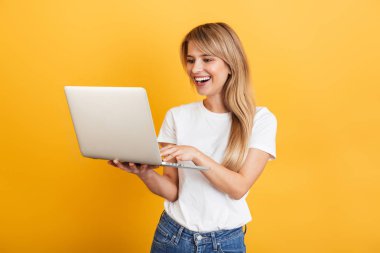 Pozitif iyimser duygusal genç sarışın kadın dizüstü bilgisayar kullanarak beyaz rahat t-shirt giymiş sarı duvar arka plan üzerinde izole poz.