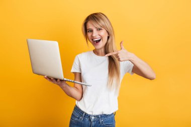 Pozitif iyimser duygusal genç sarışın kadın dizüstü bilgisayar kullanarak beyaz rahat t-shirt giymiş sarı duvar arka plan üzerinde izole poz.
