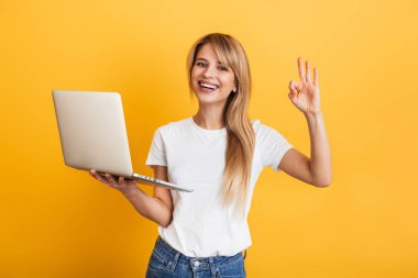 Pozitif iyimser duygusal genç sarışın kadın dizüstü bilgisayar kullanarak beyaz rahat t-shirt giymiş sarı duvar arka plan üzerinde izole poz.