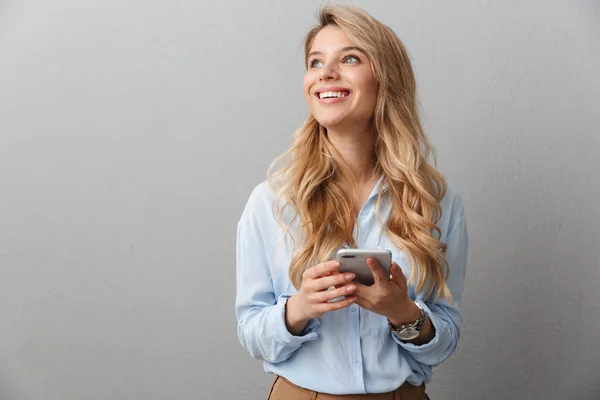 Gelukkig jonge mooie blonde zakenvrouw poseren geïsoleerde grijze muur achtergrond met behulp van mobiele telefoon. — Stockfoto