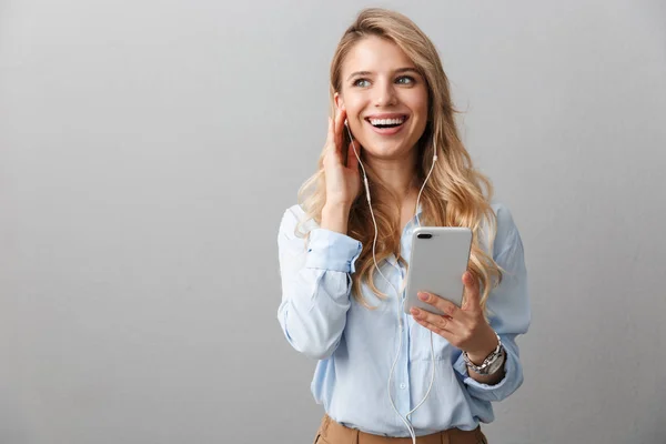 Gelukkig jonge mooie blonde zakenvrouw poseren geïsoleerde grijze muur achtergrond met behulp van mobiele telefoon luisteren muziek met oortelefoons. — Stockfoto