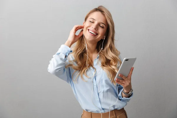 Gelukkig jonge mooie blonde zakenvrouw poseren geïsoleerde grijze muur achtergrond met behulp van mobiele telefoon luisteren muziek met oortelefoons. — Stockfoto