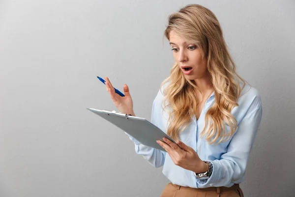 Geschokt jonge mooie blonde zakenvrouw poseren geïsoleerde grijze muur achtergrond Holding Klembord. — Stockfoto