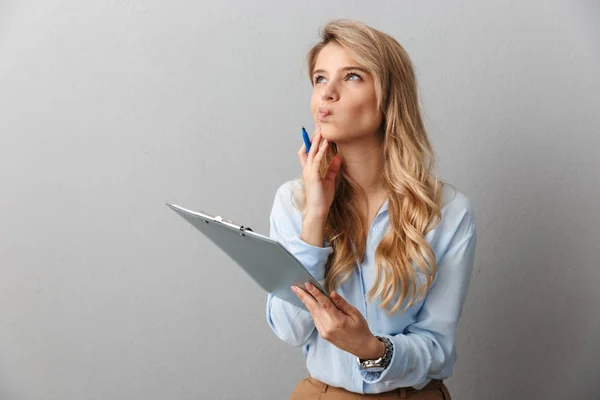 Thinking young pretty blonde business woman posing isolated grey wall background holding clipboard. — Stock Photo, Image