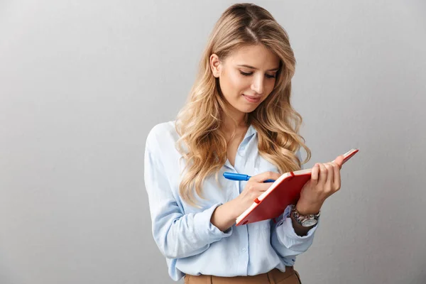 Glücklich junge hübsche blonde Geschäftsfrau posiert isoliert graue Wand Hintergrund schreiben Notizen in Notizbuch. — Stockfoto