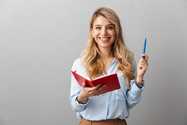 Gelukkig jonge mooie blonde zakenvrouw poseren geïsoleerde grijze muur achtergrond schrijven notities in notebook. — Stockfoto