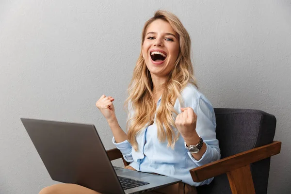 Šťastná mladá pěkná blondýnka obchodní žena, která si na pozadí izolované šedé zdi dělá gesto pro vítěze pomocí přenosného počítače. — Stock fotografie