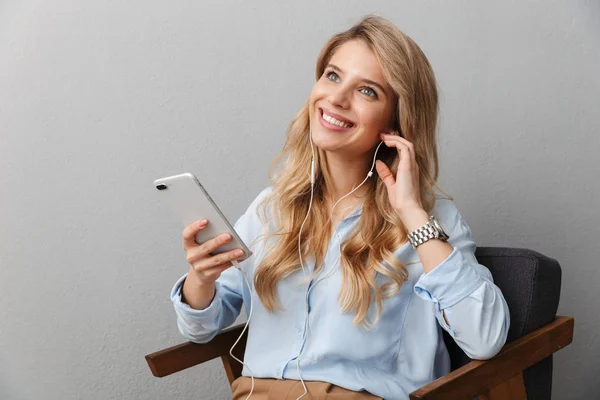 Boldog fiatal csinos szőke üzleti nő jelentő elszigetelt szürke fal háttér segítségével mobiltelefon hallgató zene fülhallgató. — Stock Fotó