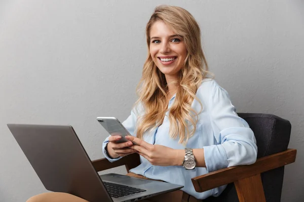 Feliz joven mujer de negocios bastante rubia posando fondo de pared gris aislado usando ordenador portátil y teléfono móvil . —  Fotos de Stock