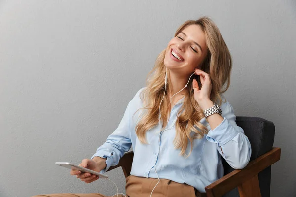 Boldog fiatal csinos szőke üzleti nő jelentő elszigetelt szürke fal háttér segítségével mobiltelefon hallgató zene fülhallgató. — Stock Fotó