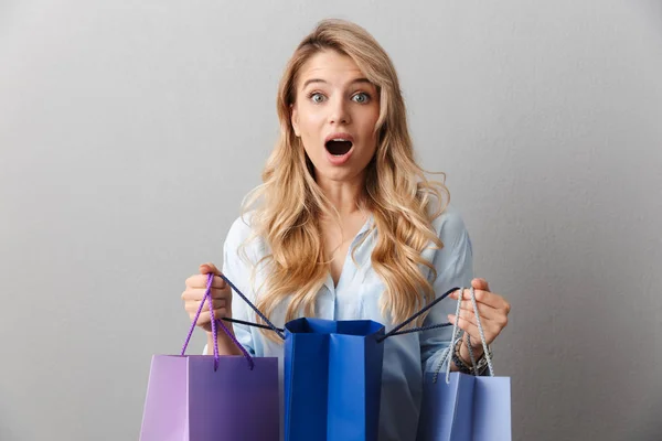 Schockiert aufgeregte junge hübsche blonde Geschäftsfrau posiert isoliert graue Wand Hintergrund mit Einkaufstaschen. — Stockfoto
