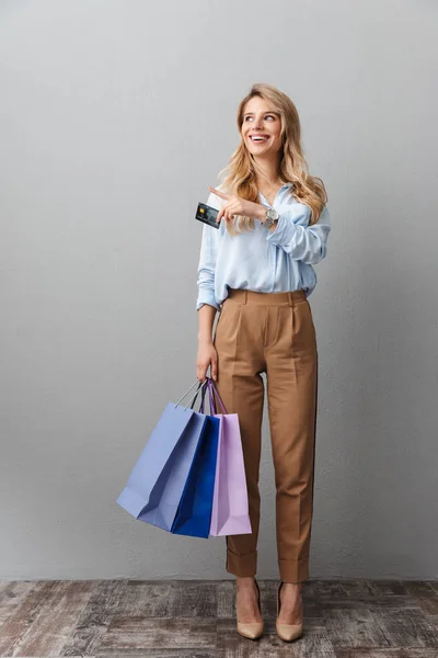 Gelukkig opgewonden jonge mooie blonde zakenvrouw poseren geïsoleerde grijze muur achtergrond Holding shopping bags. — Stockfoto