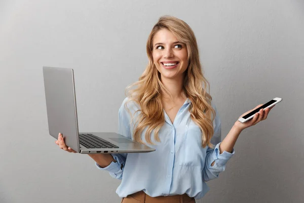 Foto de encantadora mulher loira de 20 anos vestida de camisa sorrindo enquanto segurava smartphone e laptop prata — Fotografia de Stock