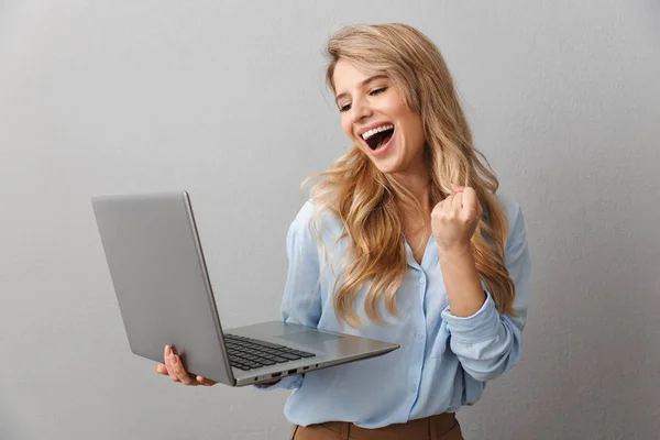 Foto de mulher loira encantada 20s vestida de camisa apertando punhos enquanto segurando e digitando no laptop prata — Fotografia de Stock