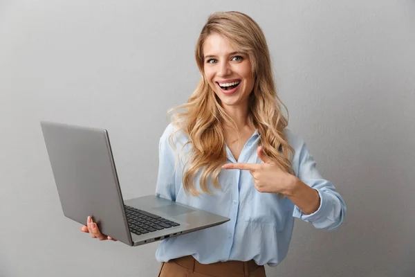 Foto de la hermosa mujer rubia de 20 años vestida con camisa sonriendo mientras sostiene y señala el dedo en el portátil de plata —  Fotos de Stock