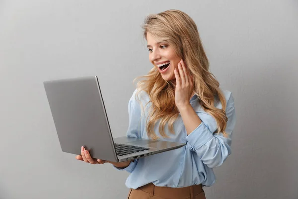 Foto de mujer rubia feliz de 20 años vestida con camisa sonriendo mientras sostiene y escribe en el portátil de plata — Foto de Stock