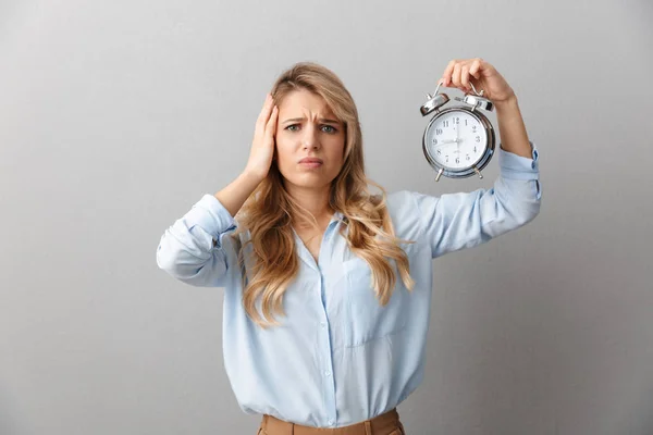Sad young pretty blonde business woman posing isolated grey wall background holding alarm clock. — Stock Photo, Image