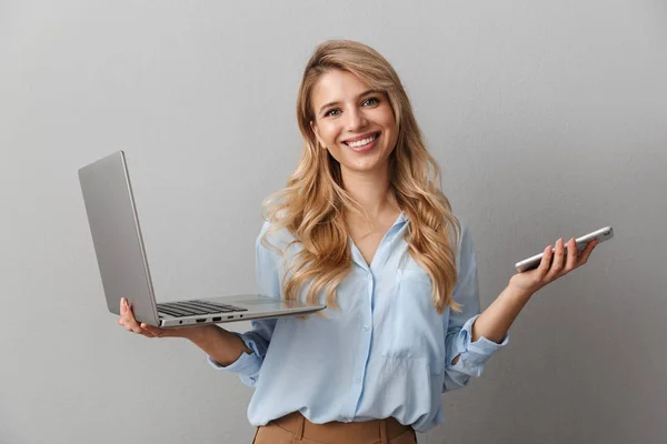Foto der zufriedenen blonden Frau 20er Jahre in Hemd lächelnd gekleidet, während sie Smartphone und silbernen Laptop in der Hand hält — Stockfoto