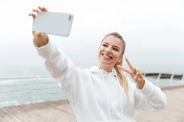 Bild einer fröhlichen netten Frau, die ein Selfie mit dem Handy macht und ein Friedenszeichen zeigt, während sie in der Nähe des Meeres spaziert — Stockfoto