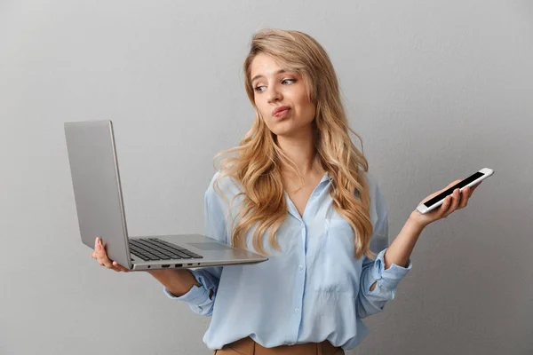 Photo of serious blonde woman 20s dressed in shirt hesitating while holding smartphone and silver laptop — Stock Photo, Image