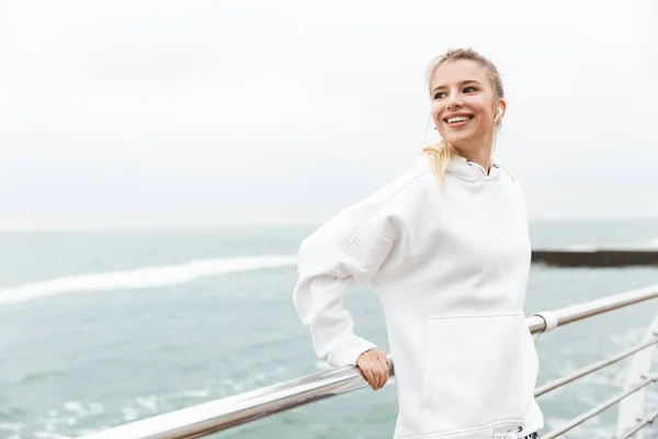 Bild der blonden fröhlichen Frau, die lächelt und Musik mit Ohrstöpseln hört, während sie in der Nähe des Meeres spaziert — Stockfoto