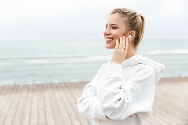 Imagem de mulher agradável feliz sorrindo e ouvindo música com fones de ouvido enquanto caminhava perto do litoral — Fotografia de Stock