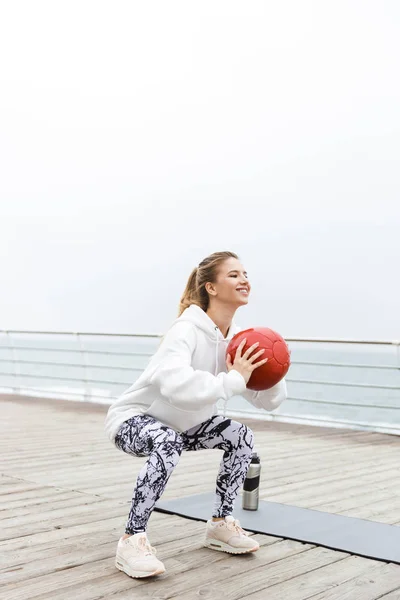 Deniz kenarında çalışırken fitness boll ile egzersiz yapan gülümseyen sportif kadın görüntü — Stok fotoğraf