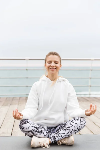 Imagem de mulher bonita sorridente meditando com dedos zen em pose de ioga no tapete de fitness enquanto trabalhava perto da beira-mar — Fotografia de Stock